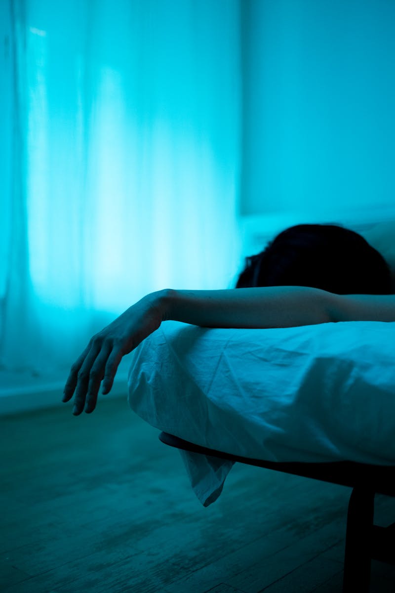 A serene and moody bedroom scene showcasing a person resting with an arm draped over the bed.