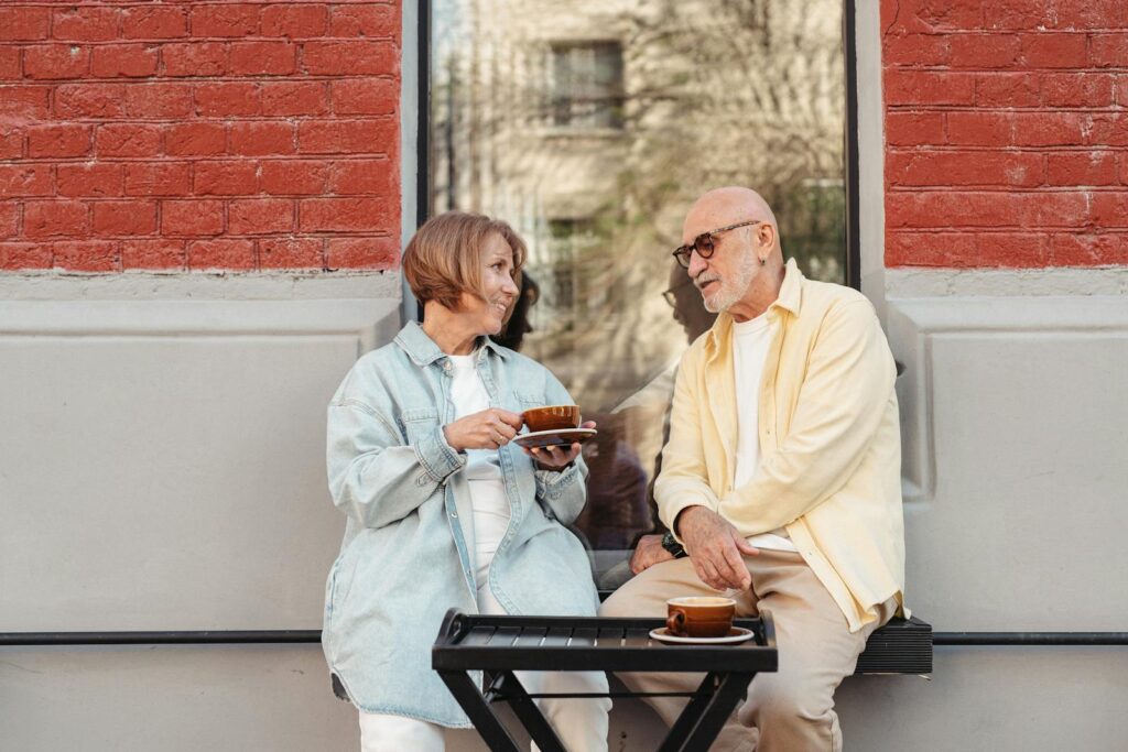 Senior couple sitting outdoors, enjoying coffee against a brick wall.