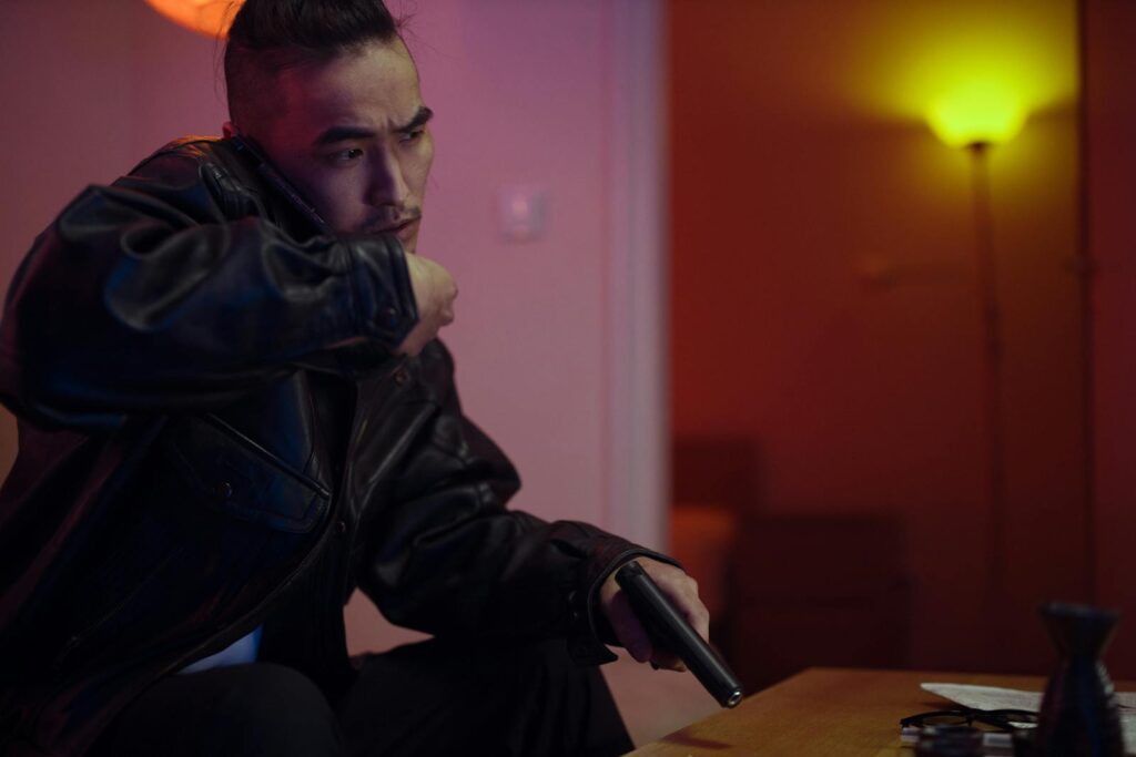 Man in a black leather jacket holding a gun while making a phone call indoors.