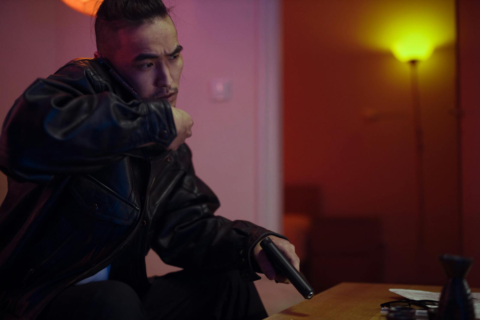 Man in a black leather jacket holding a gun while making a phone call indoors.
